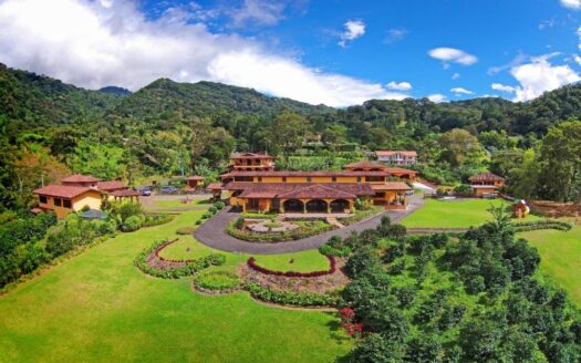 A resort with terracotta buildings arranged around curved driveways, lush gardens, and rolling green lawns set against forested hills under a blue sky.