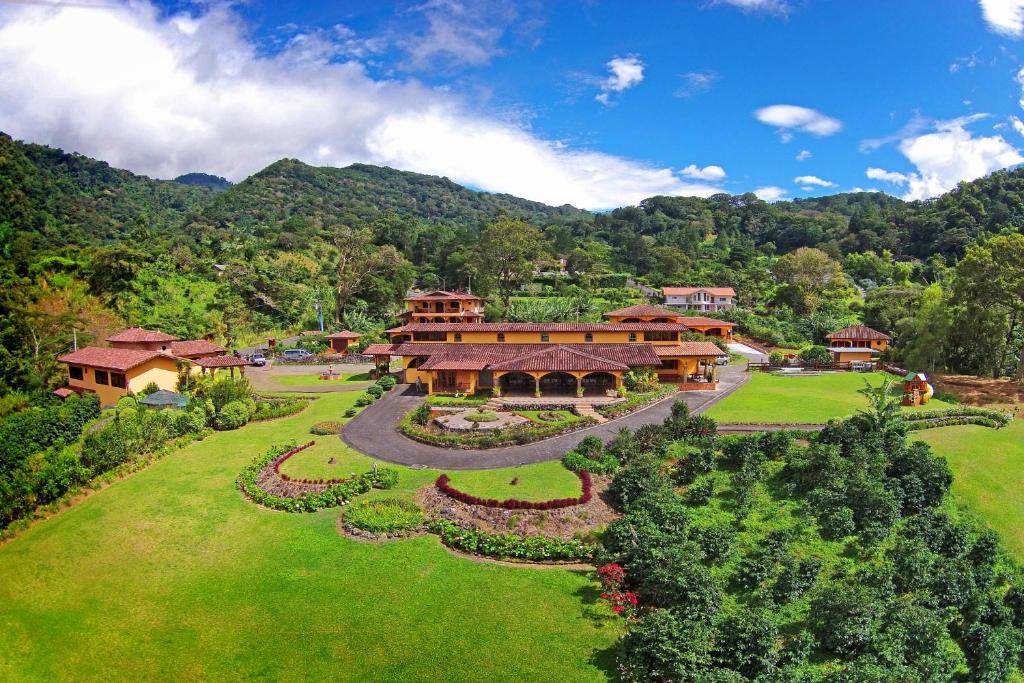 A resort with terracotta buildings arranged around curved driveways, lush gardens, and rolling green lawns set against forested hills under a blue sky.