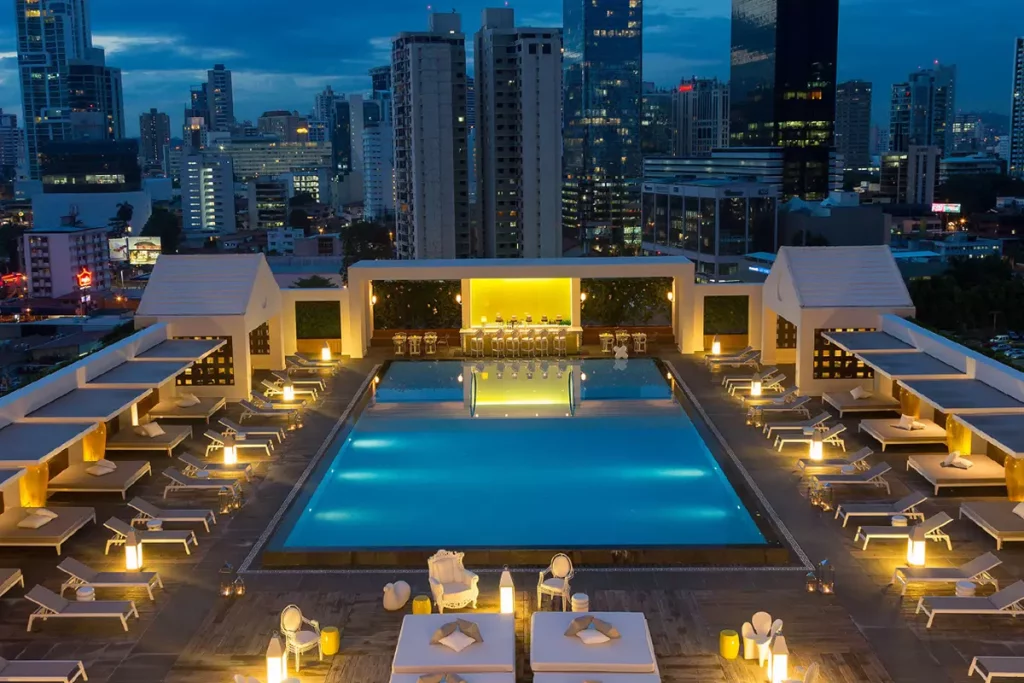 Rooftop pool area at dusk with illuminated lounge chairs along both sides and a bar at the far end, city skyline in the background