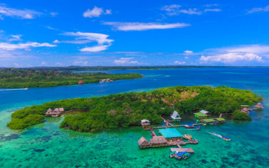 Aerial view of turquoise sea with lush green islands and overwater bungalows along a wooden jetty under a bright blue sky