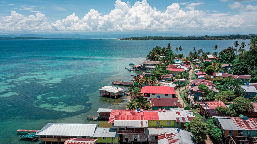 Aerial view of a tropical coastal village with colorful houses along the shore, palm trees, and turquoise water.