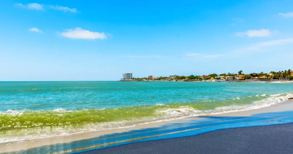 Sunny beach scene with turquoise water, white surf, and a distant coastal town along the shore.