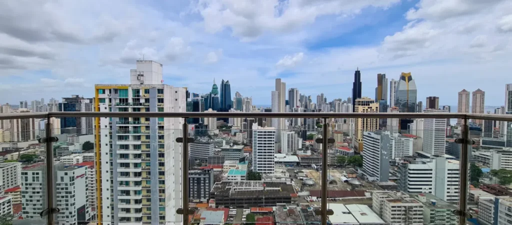 Panoramic city skyline viewed from a high balcony, with a glass railing in the foreground and numerous tall buildings under a partly cloudy sky.
