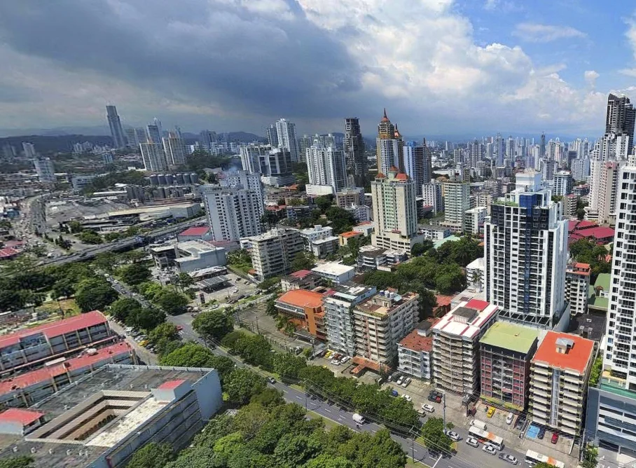 A dense urban skyline seen from above with numerous high-rise buildings, tree-lined streets, and a cloudy sky above the city.