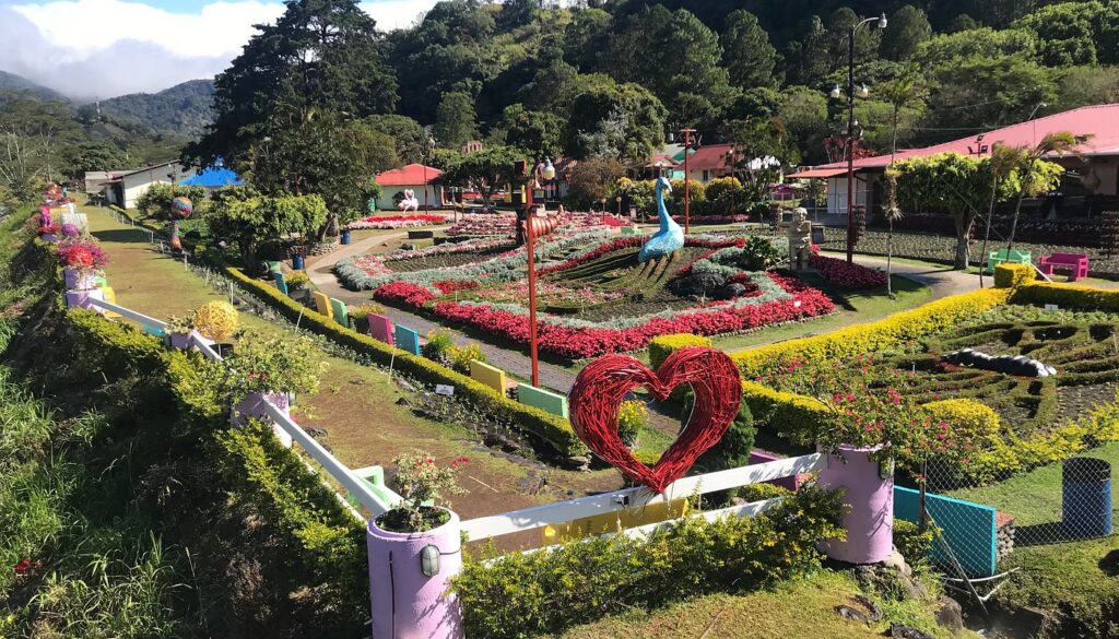 Colorful botanical garden with a woven red heart sculpture in the foreground, surrounded by blooming flower beds and decorative swan statues.
