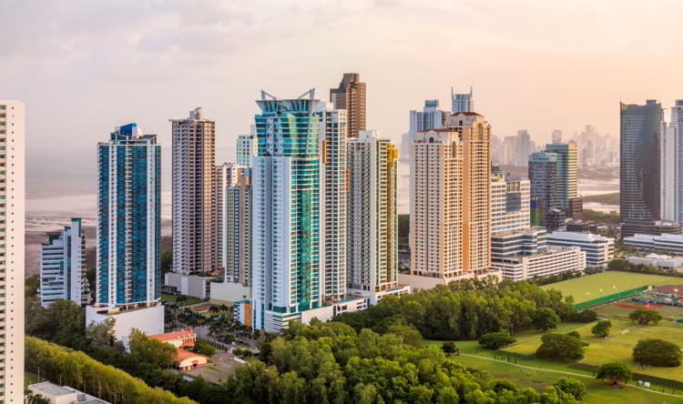 Skyline of tall glass high-rise buildings beside a green park and trees, with water visible in the distance at sunset.