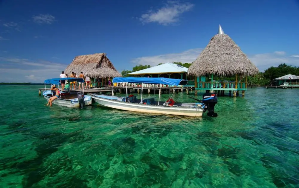 Clear tropical water with boats docked at a thatched-roof pier and huts on stilts over turquoise sea.