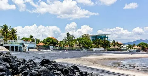 Tropical beach scene with dark rocks in the foreground, light sand, palm trees, and resort buildings along the shore under a blue sky.