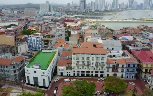 Aerial view of a coastal city with colorful mid-rise buildings near a river, a modern skyline in the distance, and red-tiled roofs in the foreground.