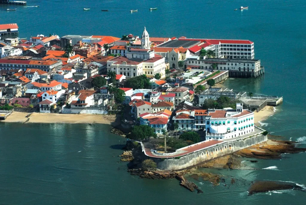 Aerial view of a seaside town with red-tiled roofs on a small island, surrounded by blue-green water.