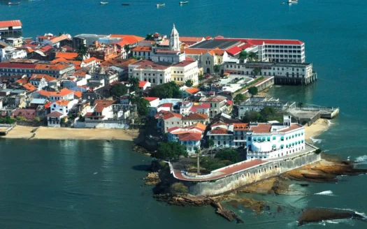 Aerial view of a seaside town with red-tiled roofs on a small island, surrounded by blue-green water.