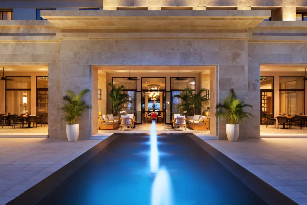 Hotel entrance with a lit central fountain pool and glass doors opening to a lounge area with seating and potted plants on each side at dusk/early evening.