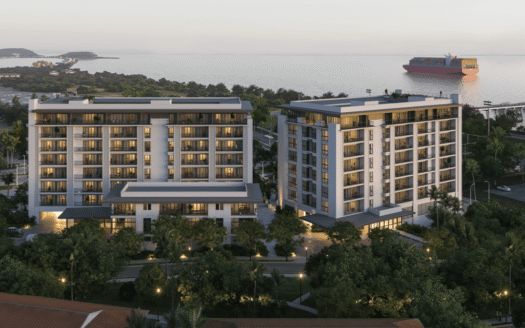 Two modern midrise residential buildings stand along the coastline with a cargo ship visible on the water in the distance.