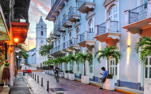 Pastel blue colonial street with arched balconies, palm planters, and a clock tower in the background at sunset.