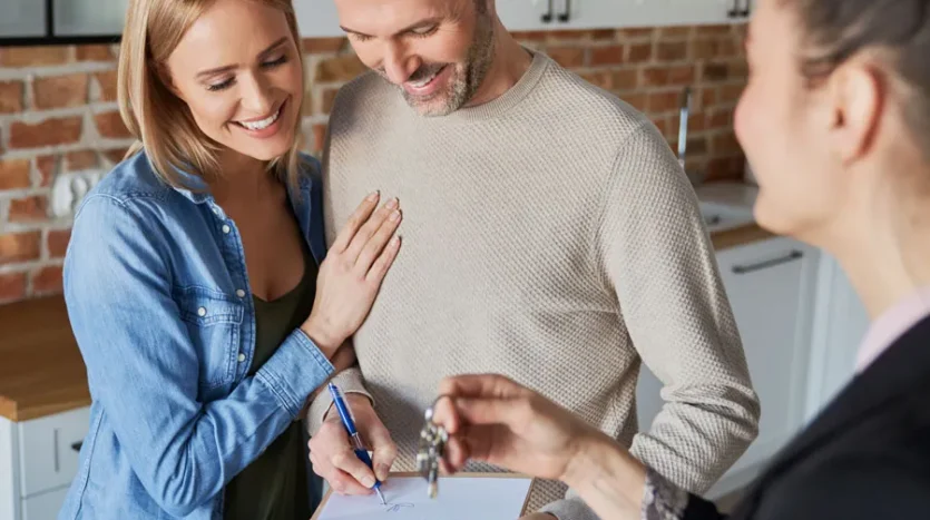 Couple in a bright kitchen sign papers with a real estate agent handing over house keys nearby.