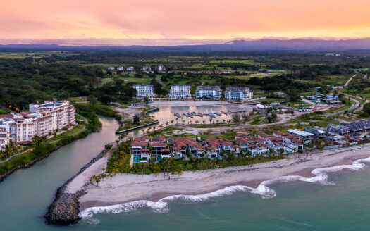 Aerial view of a seaside resort with white buildings along a sandy beach, a canal, palm trees, and a small marina at sunset.
