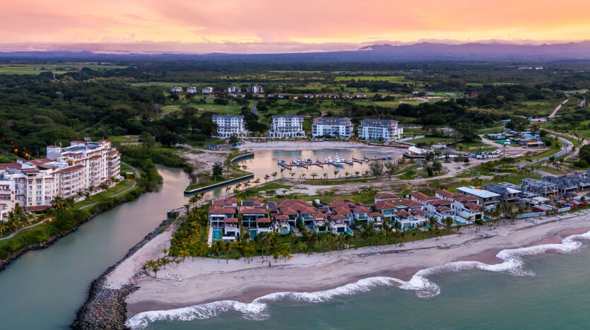 Aerial view of a seaside resort with white buildings along a sandy beach, a canal, palm trees, and a small marina at sunset.
