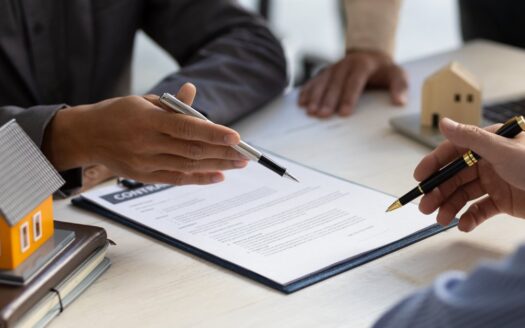 Two people sign a real estate contract on a desk, with a model orange house and pens nearby.