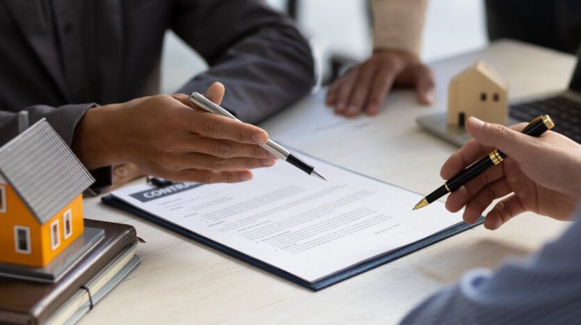 Two people sign a real estate contract on a desk, with a model orange house and pens nearby.