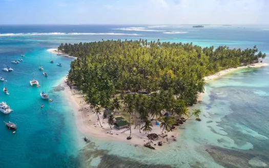 Sell-Prroperty-in-Panama-Tax - Panama Elite Homes A tropical island densely lined with palm trees, surrounded by turquoise water and anchored boats along a sandy shoreline.