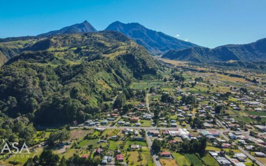 Aerial view of a valley town nestled among green hills and a distant mountain range under a bright blue sky (CASA watermark in the lower-left corner).