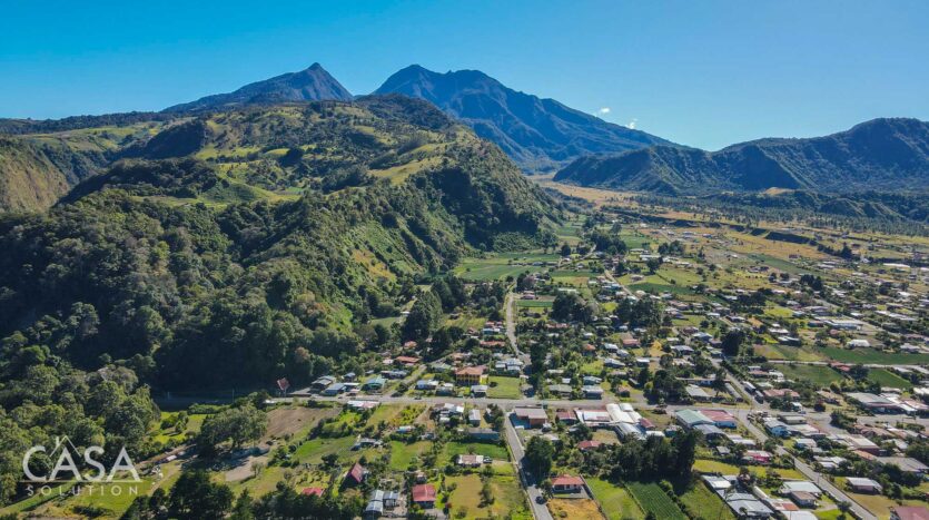 Aerial view of a valley town nestled among green hills and a distant mountain range under a bright blue sky (CASA watermark in the lower-left corner).