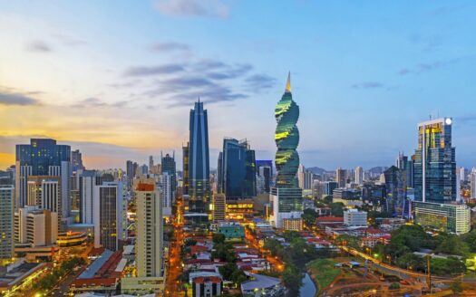 Dusk skyline of a modern city with glass skyscrapers, including a spiraling green tower and illuminated streets.