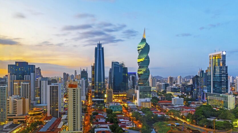 Dusk skyline of a modern city with glass skyscrapers, including a spiraling green tower and illuminated streets.