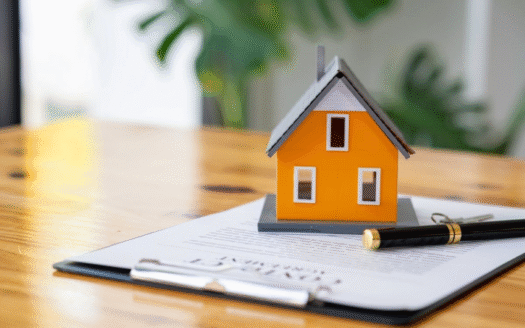 Orange miniature house sits on top of a clipboard with a document and a pen, on a wooden table (real estate or home loan context).