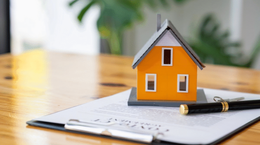 Orange miniature house sits on top of a clipboard with a document and a pen, on a wooden table (real estate or home loan context).