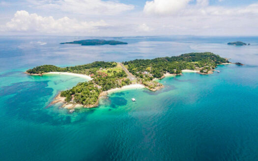 Aerial view of a tropical island connected by a road bridge, with turquoise lagoons, white-sand beaches, and lush green forests.