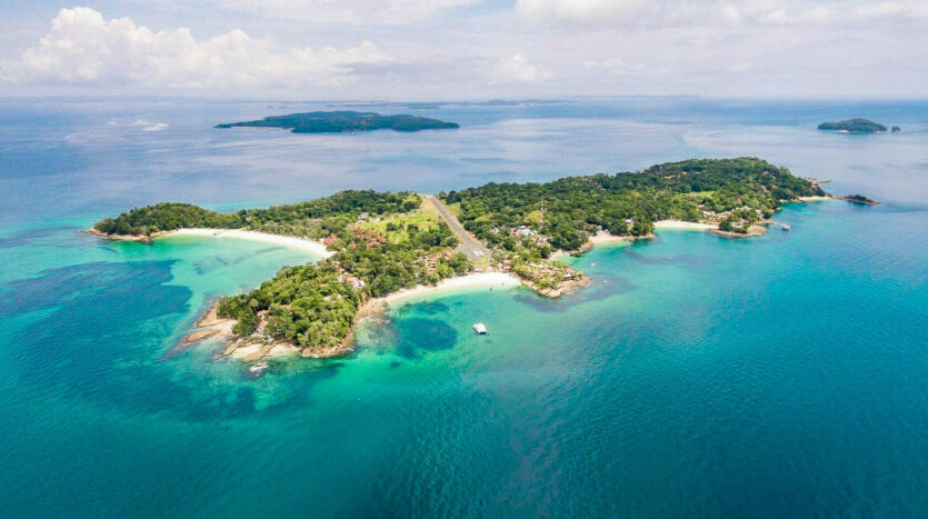 Aerial view of a tropical island connected by a road bridge, with turquoise lagoons, white-sand beaches, and lush green forests.