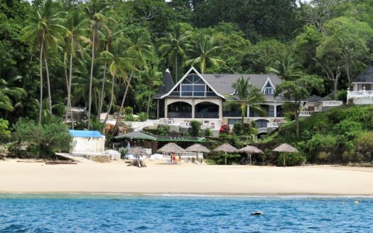 Tropical beach scene with a large beachfront villa framed by palm trees, white railings, and thatched shade structures on the sand, calm turquoise water in the foreground.