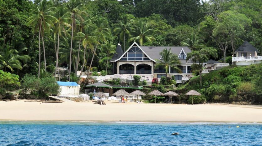 Tropical beach scene with a large beachfront villa framed by palm trees, white railings, and thatched shade structures on the sand, calm turquoise water in the foreground.