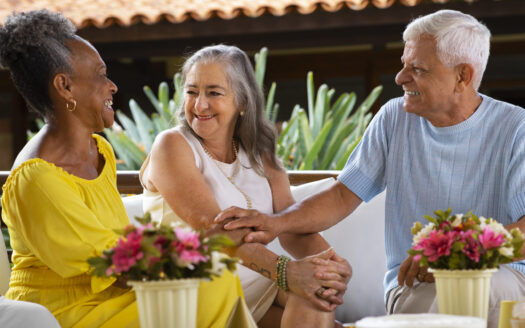 Three smiling seniors sit at an outdoor table, holding hands and chatting, with flower pots in the foreground.