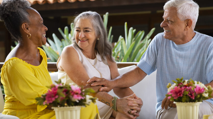 Three smiling seniors sit at an outdoor table, holding hands and chatting, with flower pots in the foreground.