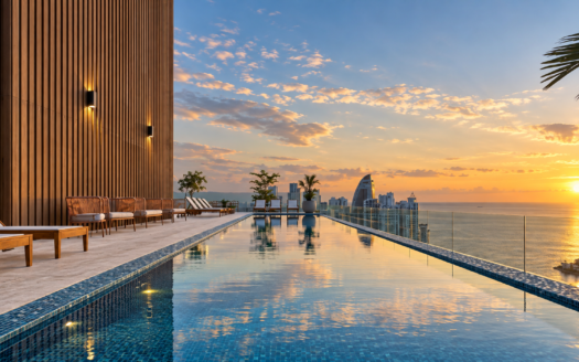 Rooftop infinity pool at sunset, with lounge chairs along a wooden wall and a distant city skyline.