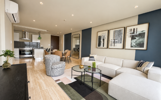 Open-plan living room with a beige sectional sofa, glass coffee tables, and a dark blue accent wall displaying three framed artworks. Kitchen visible in the background.