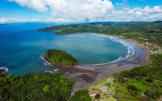 Aerial view of a curved sandy beach along a lush green hillside with blue ocean and a small coastal village.