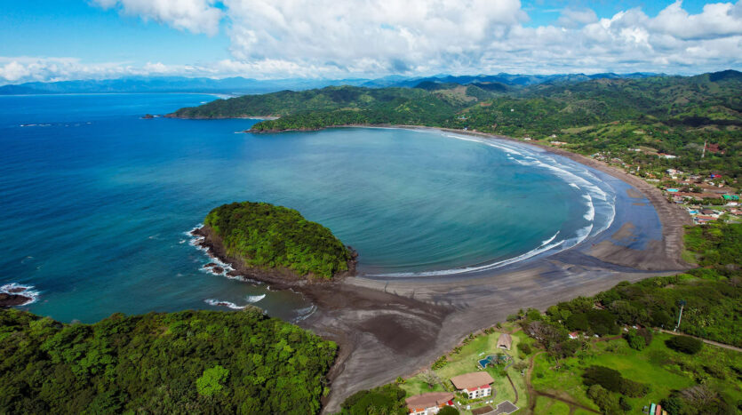 Aerial view of a curved sandy beach along a lush green hillside with blue ocean and a small coastal village.